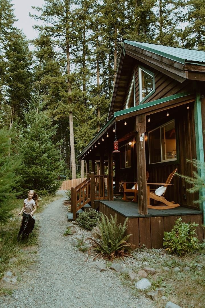 This may contain: a woman walking down a dirt road next to a wooden cabin with lights on it