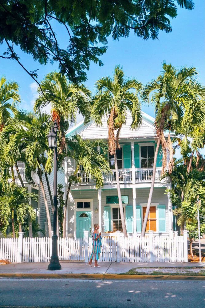 This may contain: a woman walking down the street in front of a blue and white house with palm trees