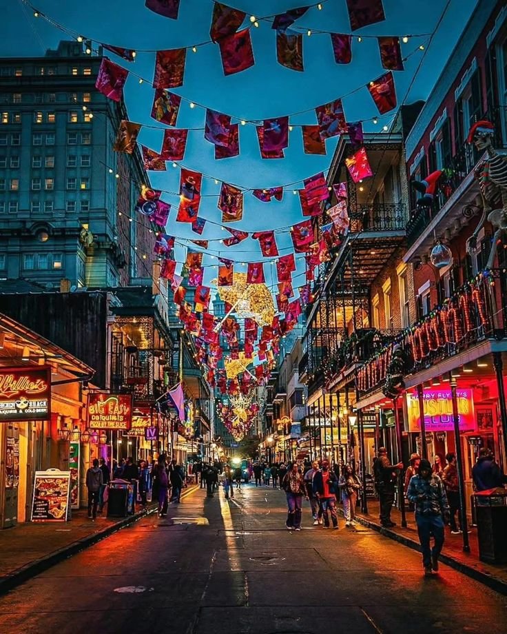 This may contain: people are walking down the street in front of buildings with flags hanging from them and lights strung above