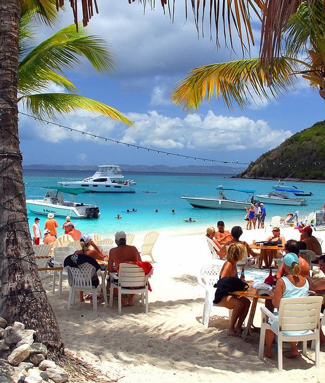 This may contain: many people are sitting at tables on the beach with boats in the water and palm trees