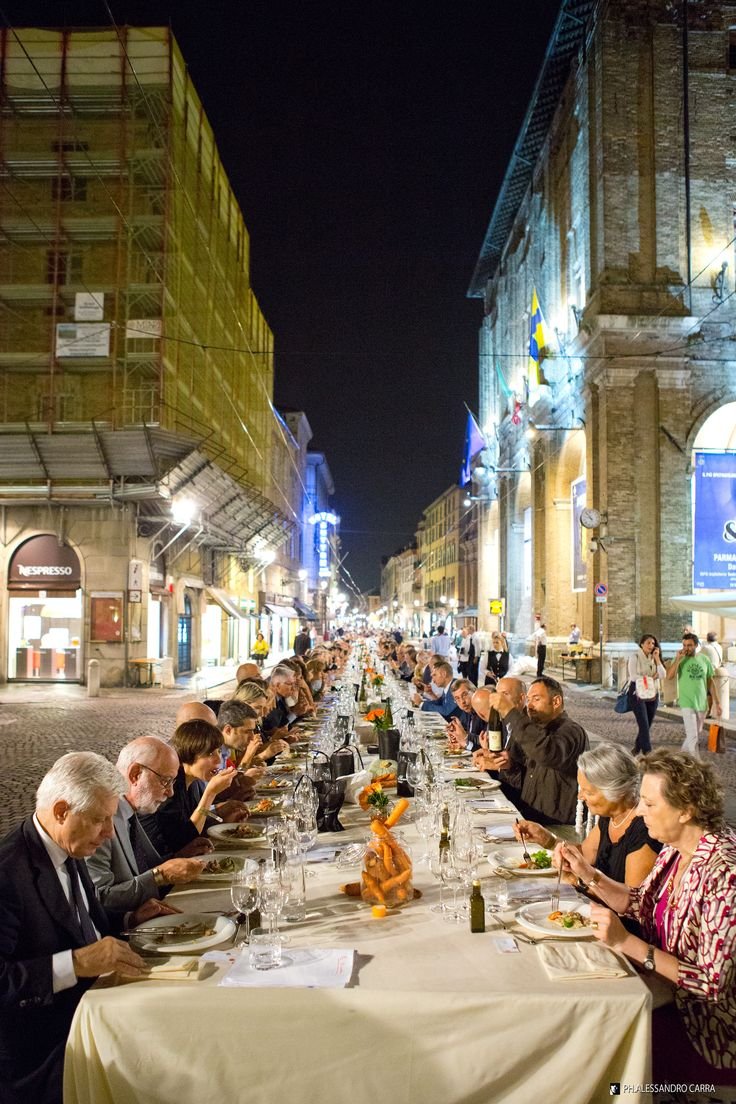 This may contain: a group of people sitting at a long table in front of tall buildings on a city street