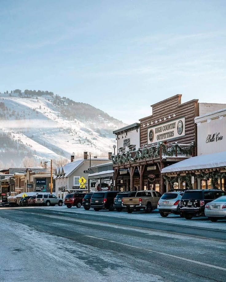 This may contain: cars parked on the side of a road in front of buildings and snow covered mountains