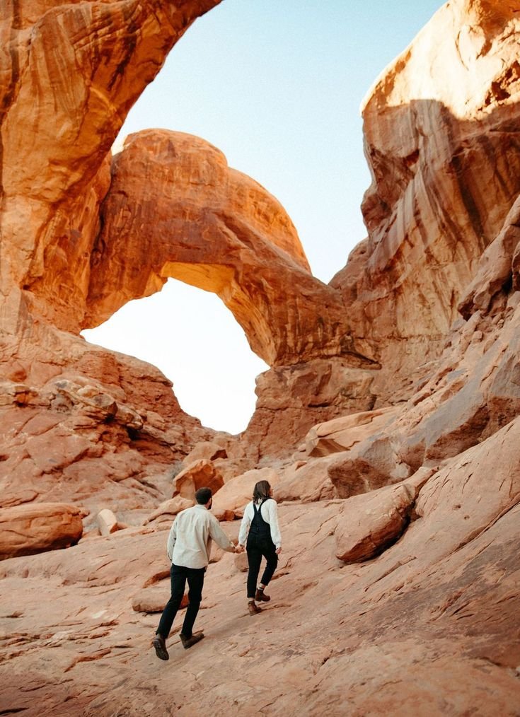 This may contain: two people are walking through the desert near large rocks and an arch in the background