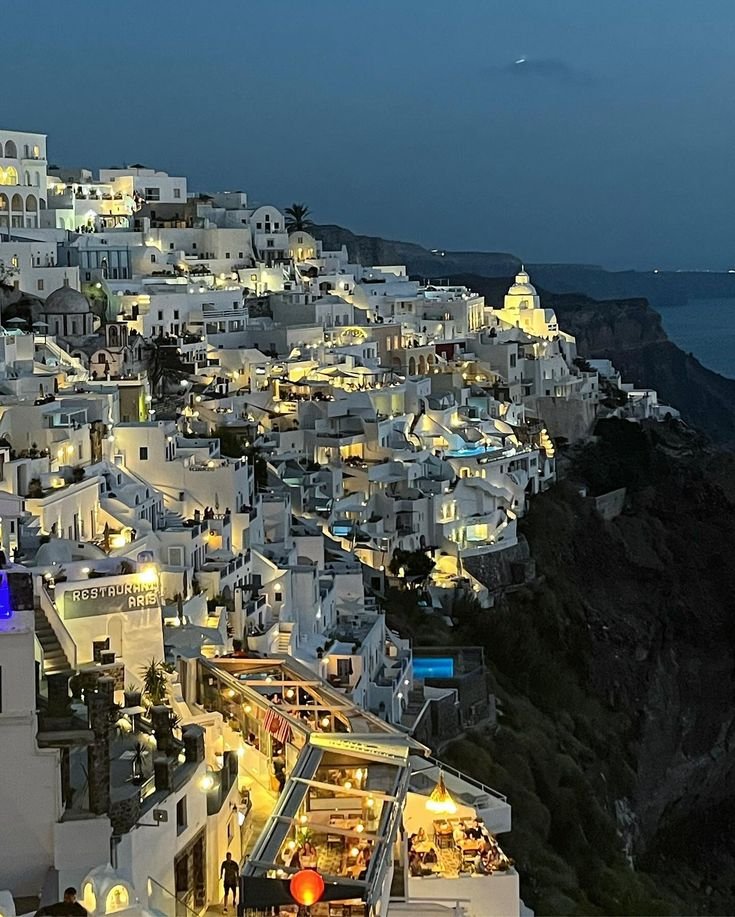 This may contain: the white buildings are lit up at night on the cliff above the town's water
