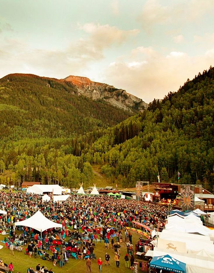 This may contain: a large group of people standing around tents in the grass with mountains in the background