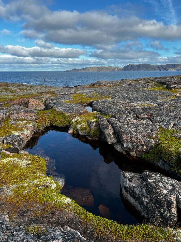 This may contain: a small pond in the middle of some rocks by the ocean with clouds and blue sky
