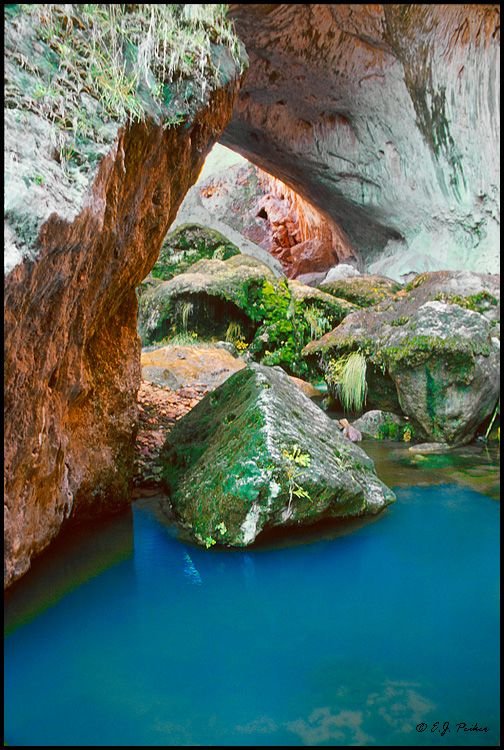 This may contain: the blue pool is surrounded by large rocks and green mossy vegetation, with an arch in the middle