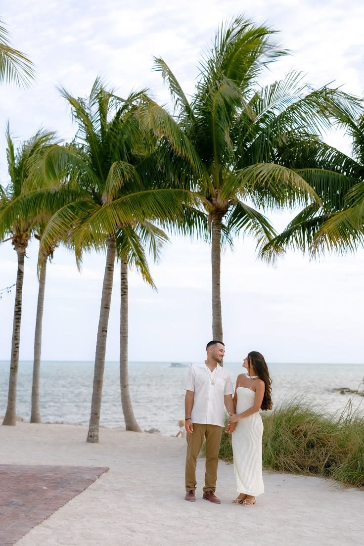 This may contain: a man and woman standing next to each other on a beach near the ocean with palm trees