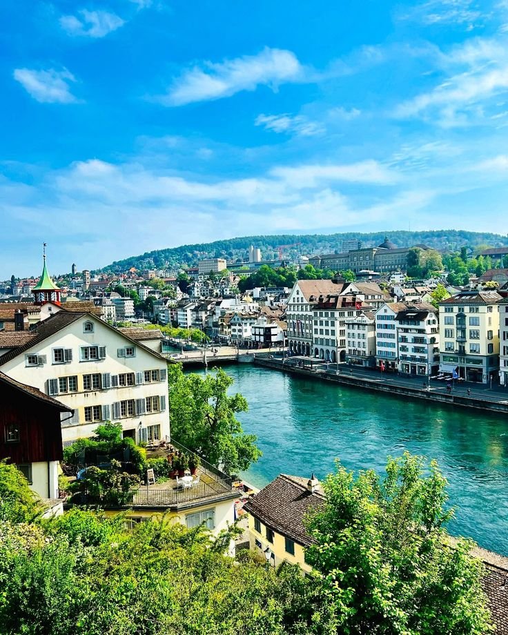 This may contain: a river running through a city next to a lush green hillside covered in lots of trees