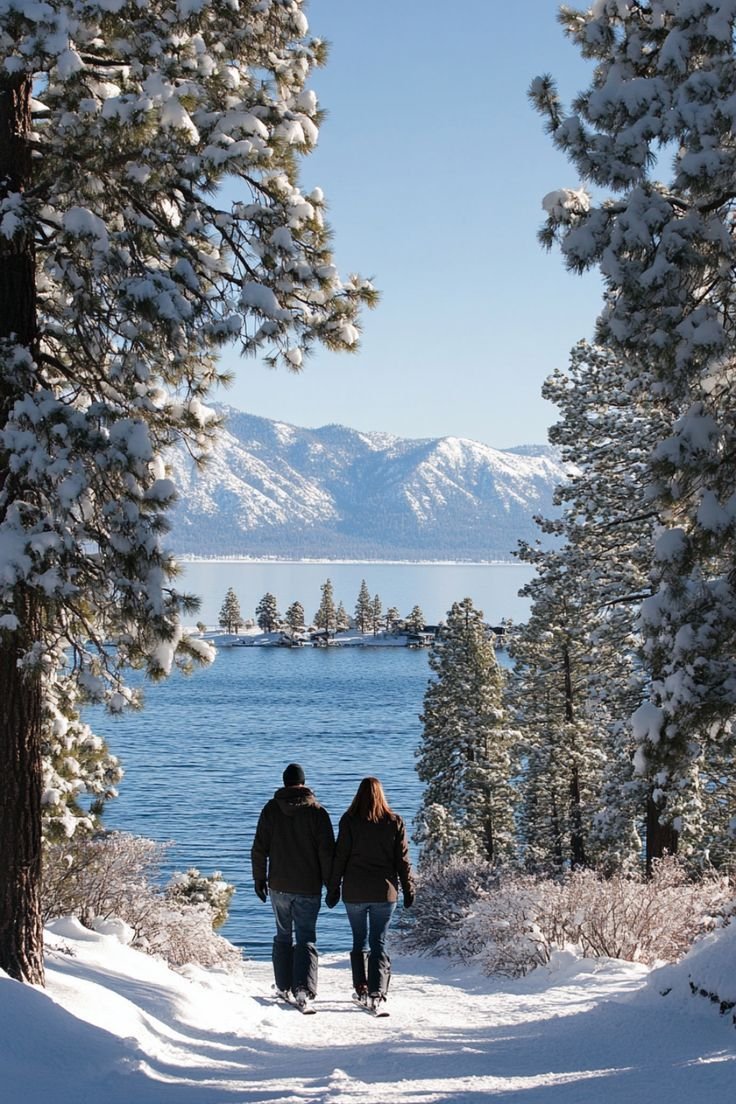 This may contain: two people walking down a snow covered path near the water's edge with trees in the foreground