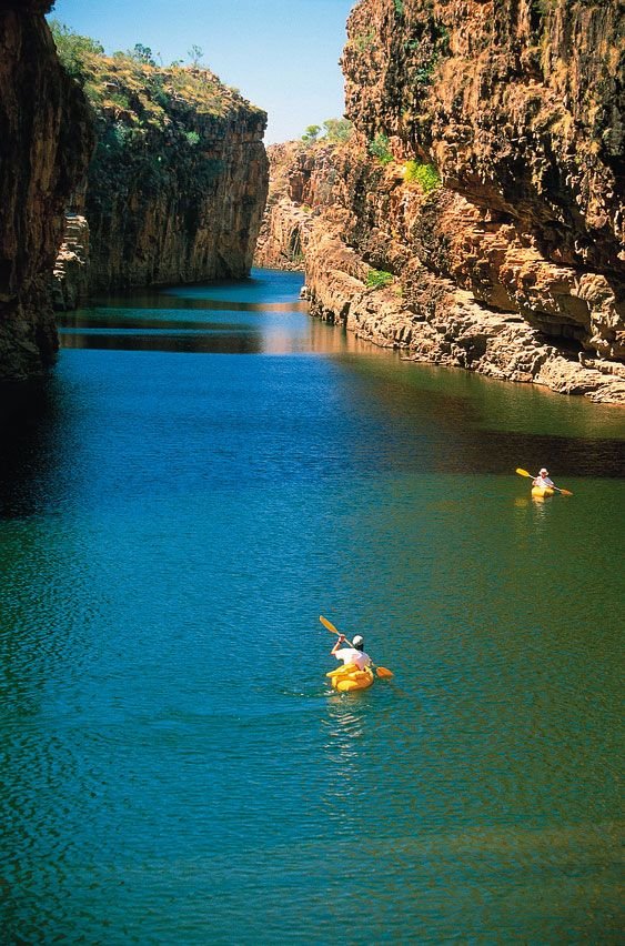 This may contain: two people in kayaks paddling through the water near some rocks and cliffs on a sunny day