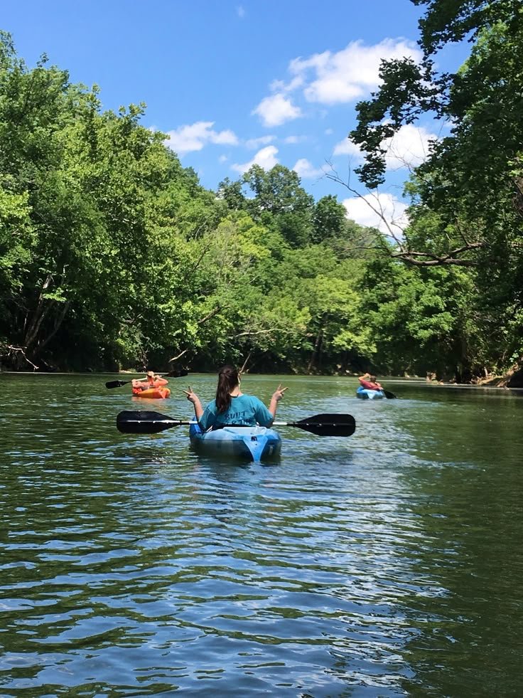 This may contain: two people in kayaks paddling down a river surrounded by green trees and blue sky