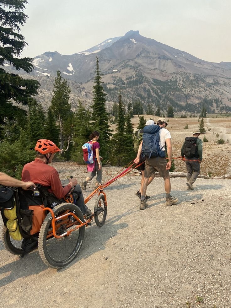 This may contain: a man in a wheel chair being pulled by two people on a mountain trail with mountains in the background