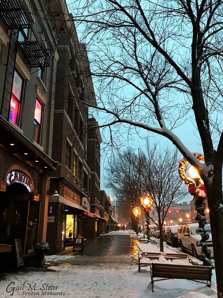 This may contain: a snowy street lined with buildings and benches