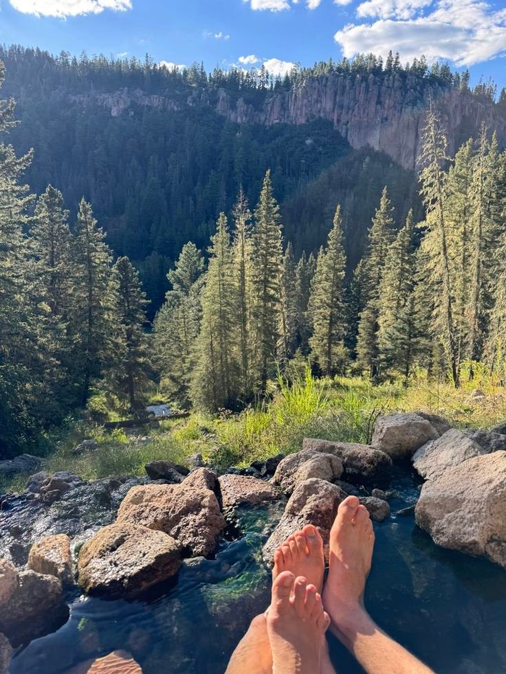 This may contain: two people are laying on rocks in the water near some trees and mountains with a river running through them