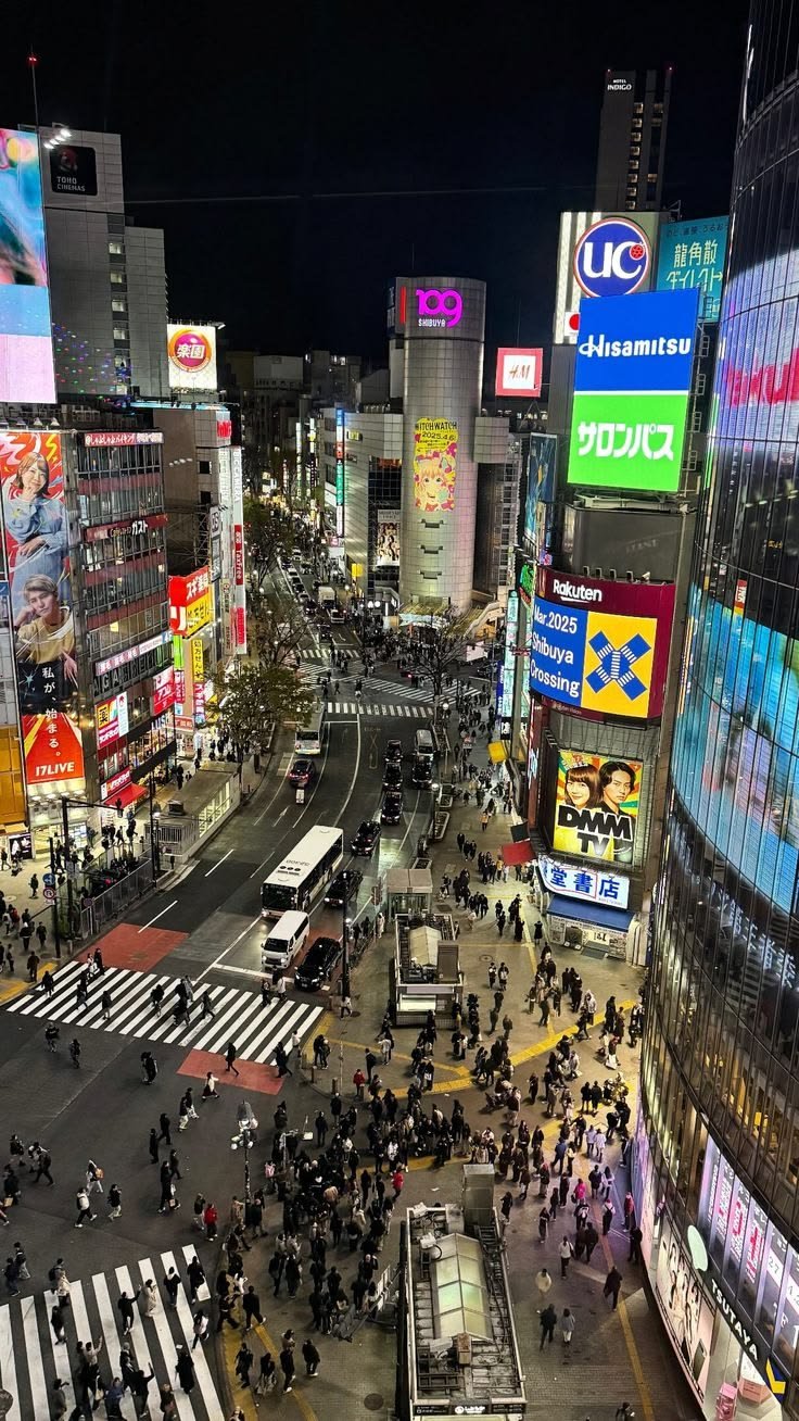 This may contain: a busy city street at night with lots of people crossing the street in front of tall buildings