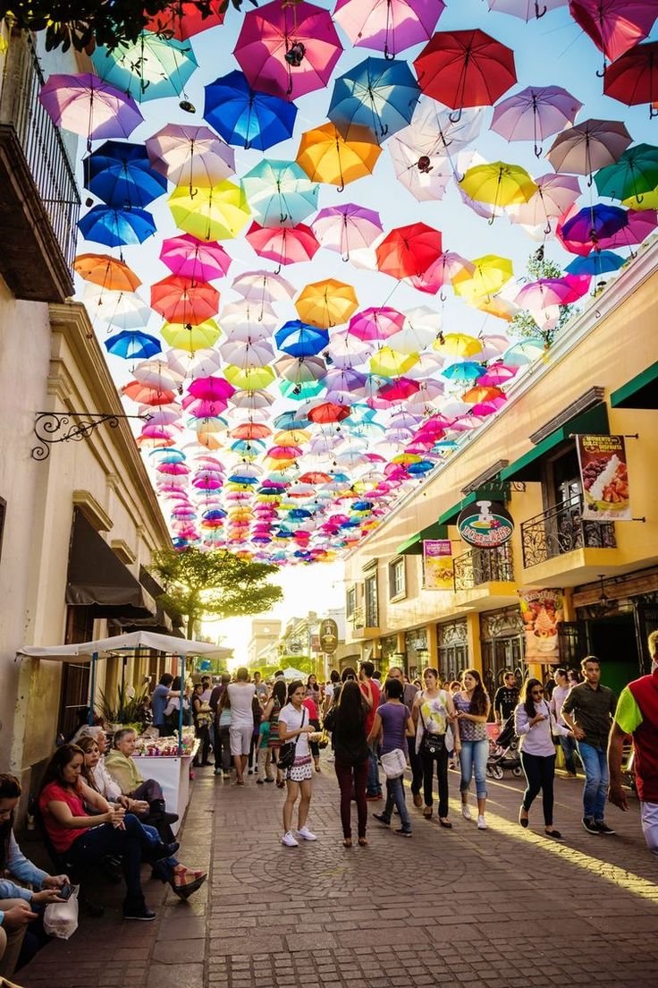 This may contain: many colorful umbrellas hanging from the ceiling in an open air market area with people walking around