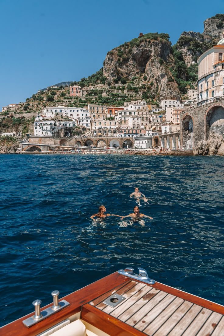 This may contain: two people are swimming in the water near some buildings and cliffs, while another person is on a boat