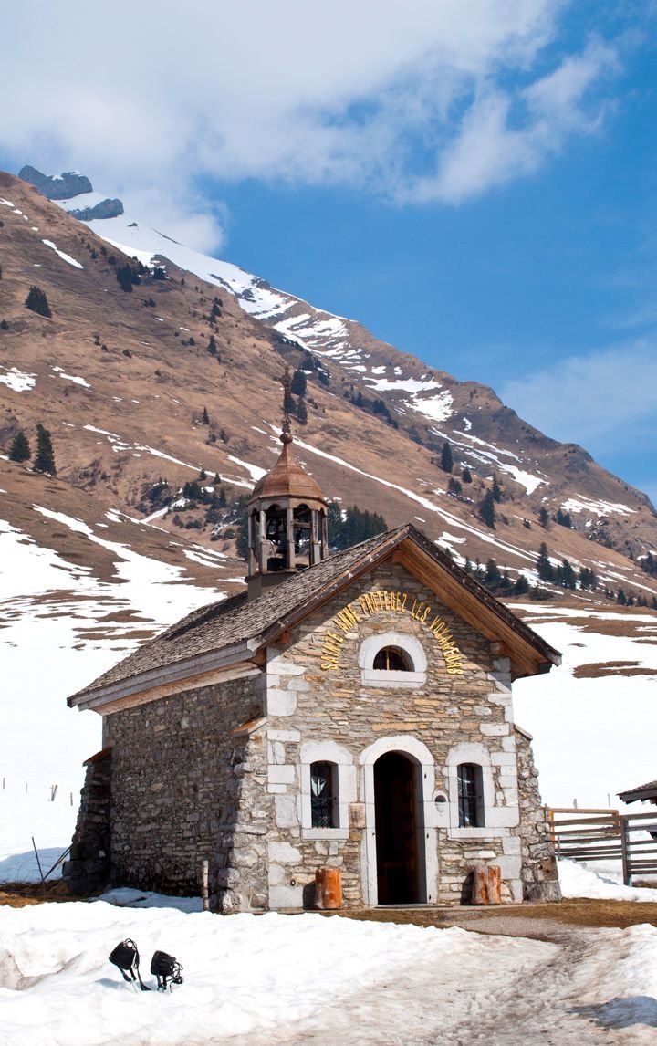 This may contain: an old stone church in the middle of winter with snow on the ground and mountains behind it