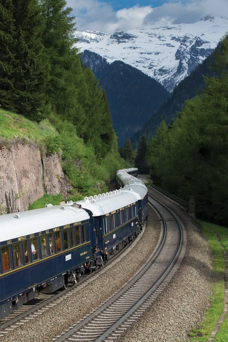 This may contain: a train traveling down tracks next to a lush green hillside covered in snow capped mountains