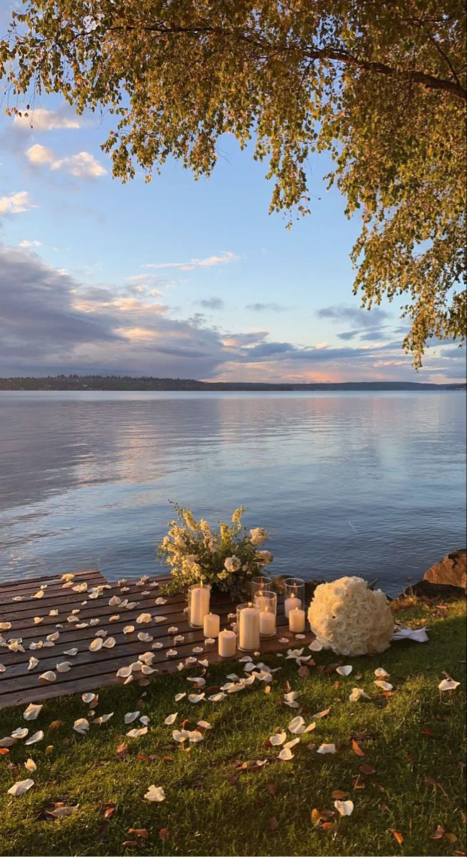 This may contain: an outdoor wedding setup with candles and flowers on the grass by the water at sunset