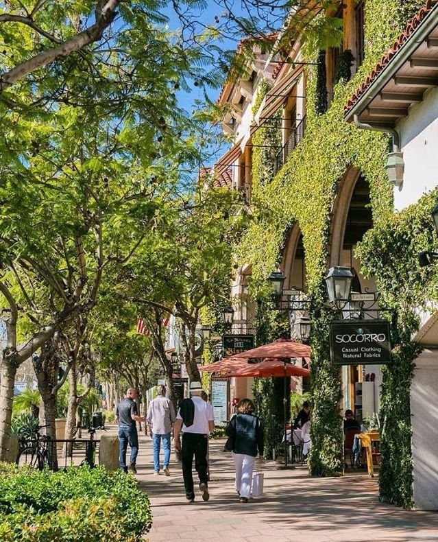 This may contain: people are walking down the sidewalk in front of buildings with trees and bushes on either side
