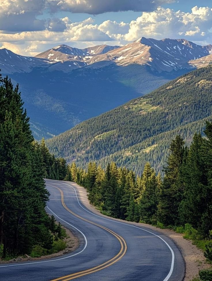 This may contain: an empty road in the middle of mountains with trees on both sides and snow capped peaks behind it
