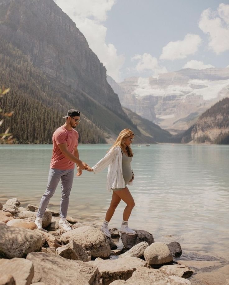 This may contain: a man and woman are holding hands by the water's edge, with mountains in the background
