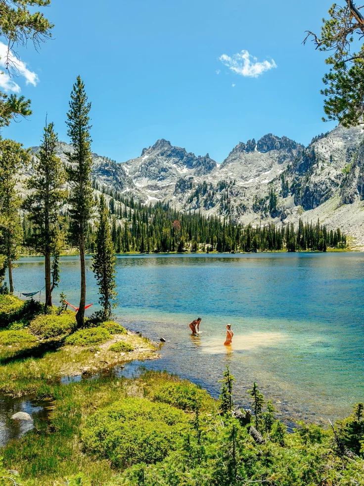 This may contain: two people are wading in the water near some trees and mountain peaks on a sunny day
