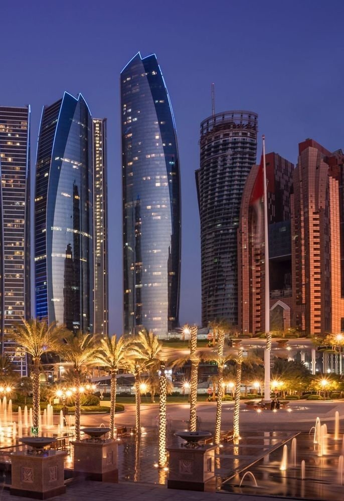 This may contain: the city is lit up at night with tall buildings in the background and palm trees
