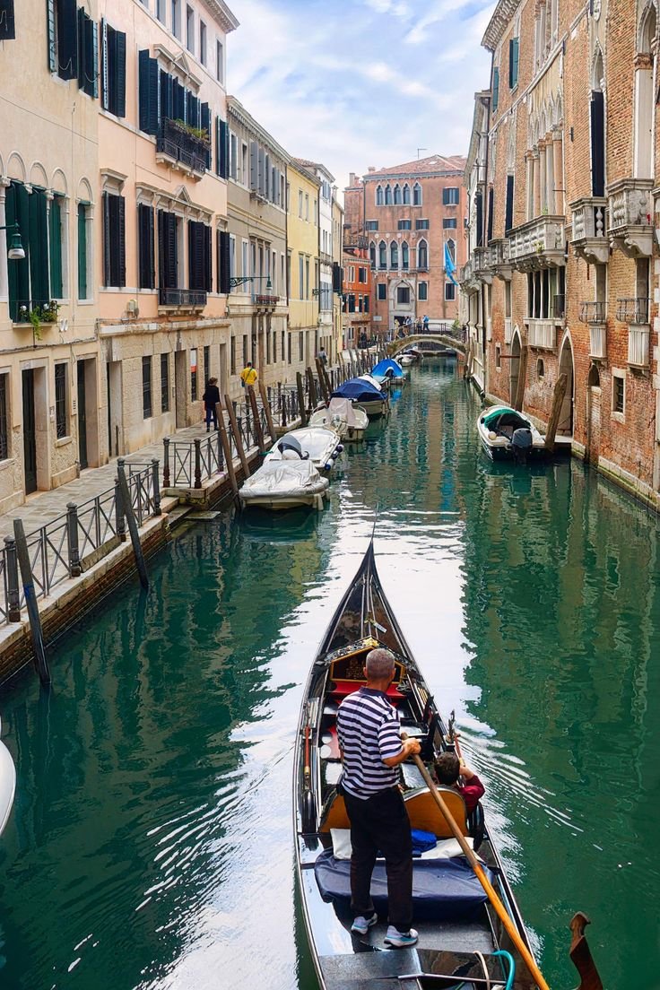 This may contain: a man standing on the side of a boat in a canal next to tall buildings