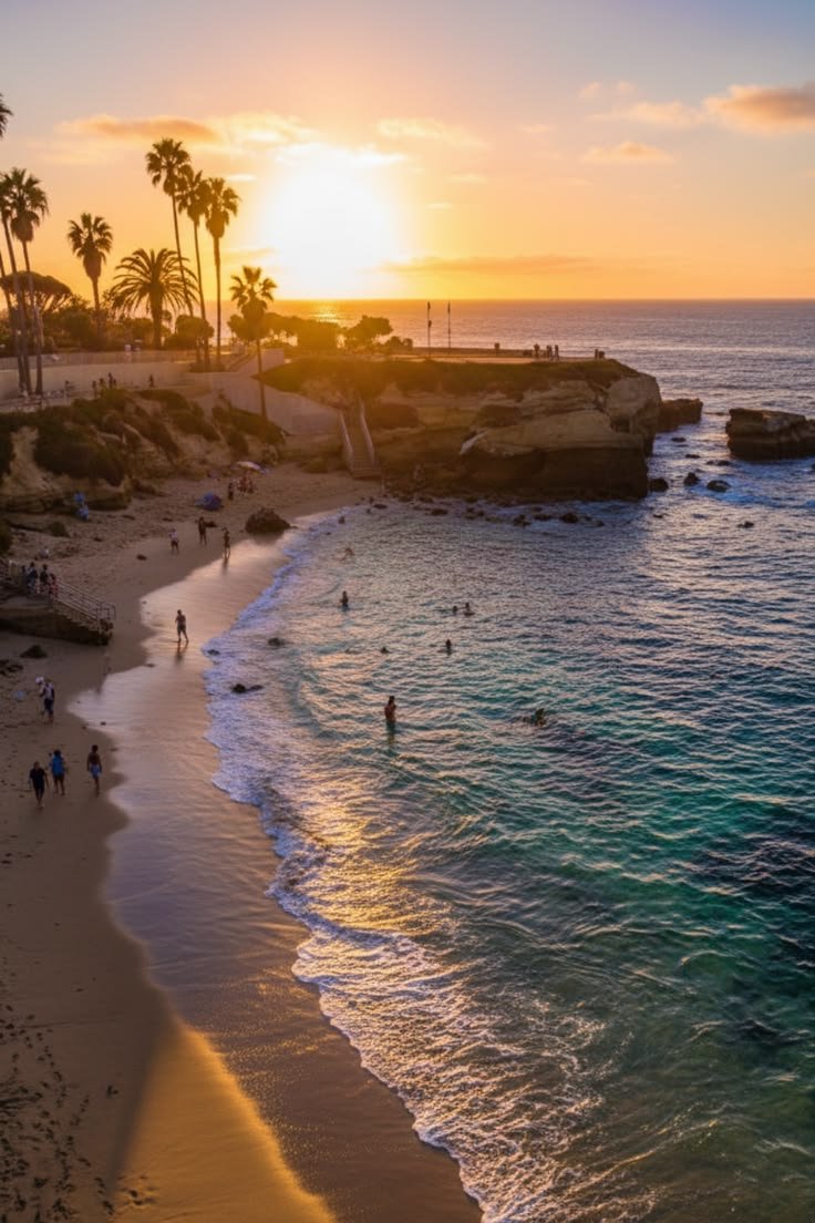 This contains: This scenic landscape captures a beautiful sunset over a sandy beach. Tall palm trees stand along the coastline during the golden hour. People are relaxing on the shore and swimming in the calm ocean water. The rocky cliffs add depth to the coastal scenery