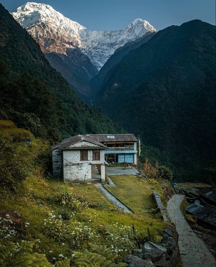 This may contain: an old house in the mountains with snow capped mountains behind it and a path leading up to it