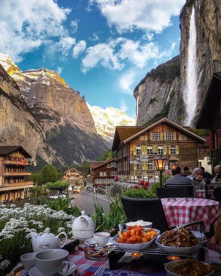 This may contain: a table with food on it in front of a mountain village and some mountains behind it