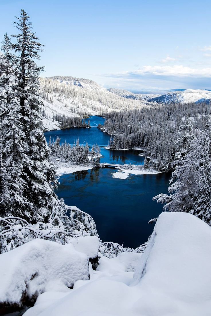 This may contain: a lake surrounded by snow covered trees in the middle of a snowy mountain range with blue water