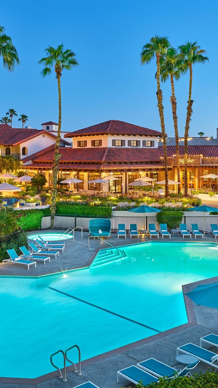 This may contain: an outdoor swimming pool with chaise lounges and palm trees in the background at dusk