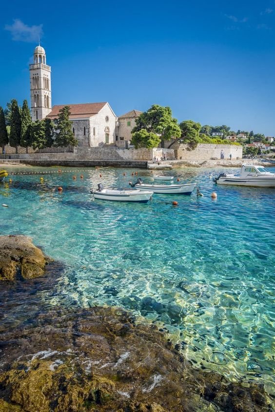 This may contain: people swimming in clear blue water next to a white church and small boats on the shore