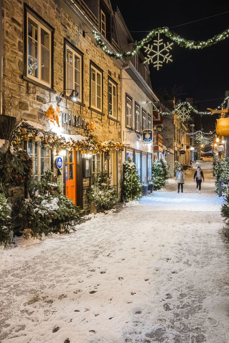 This may contain: a snowy street with people walking down it and christmas lights strung over the buildings on both sides
