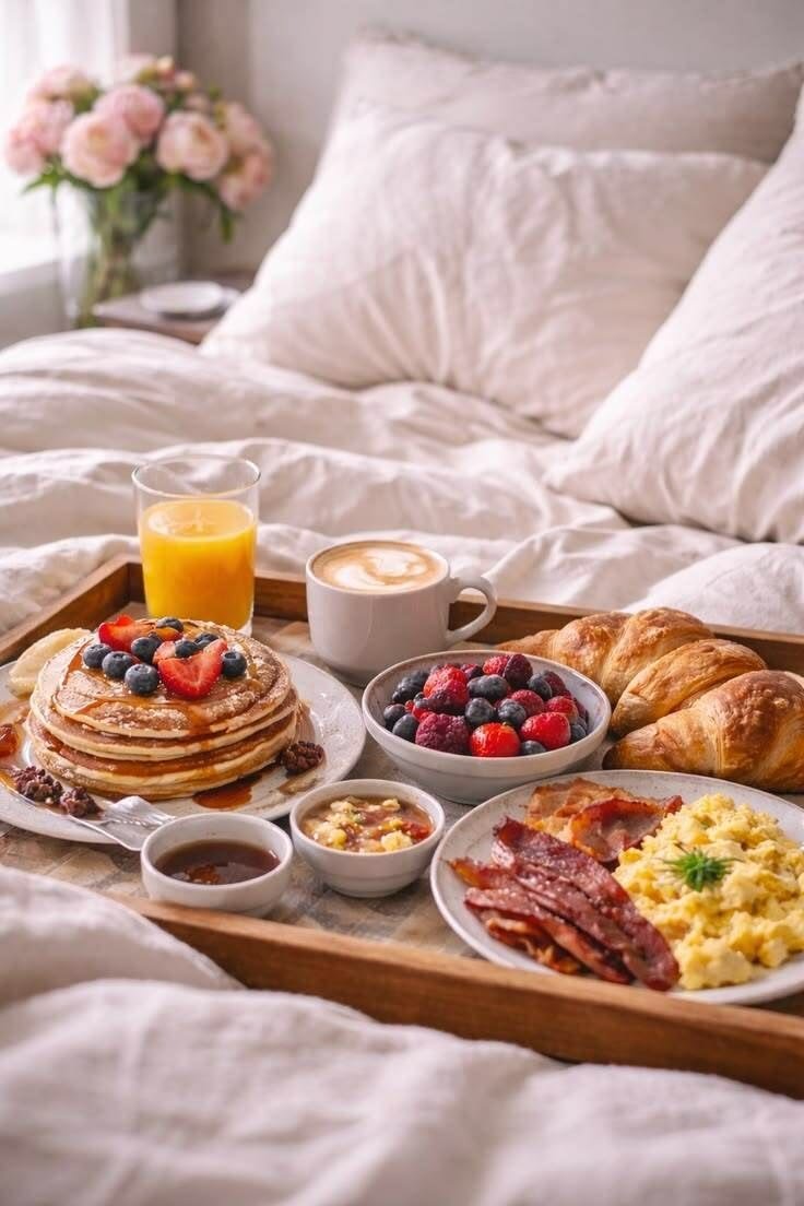 This may contain: a breakfast tray with eggs, bacon, pancakes, fruit and coffee on the bed