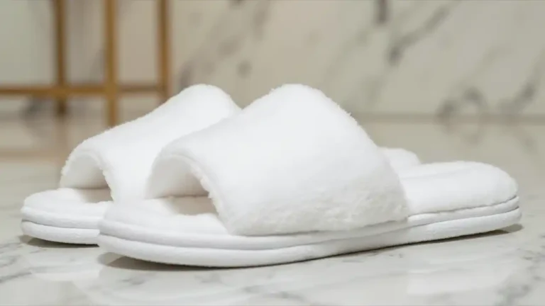 A pair of plush white hotel slippers neatly placed on a luxurious marble bathroom floor, with a folded white towel and elegant hotel amenities in the background