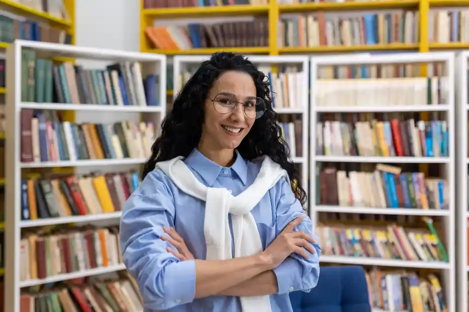 Confident female librarian standing among bookshelves for National School Librarian Day.