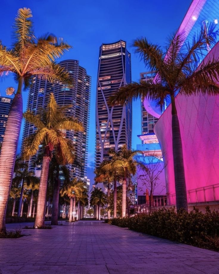 This may contain: palm trees and buildings in the city at night with bright lights on them, along side a walkway