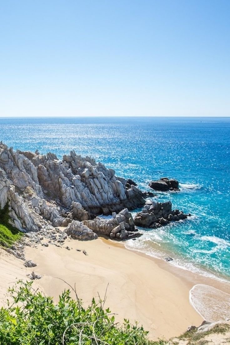 This may contain: the beach is next to some rocks and clear blue water with waves coming in on it