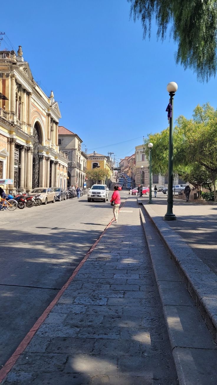 This may contain: a woman walking down the middle of a street with cars parked on both sides and buildings in the background