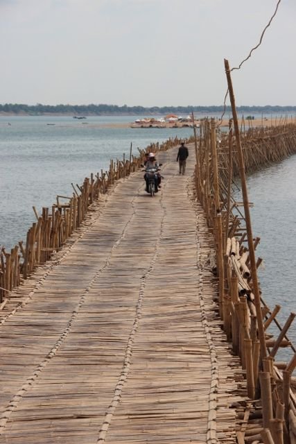 This may contain: two people walking on a wooden bridge over water with bamboo poles in the foreground