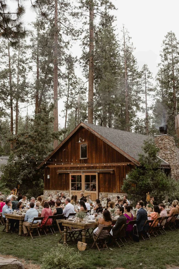 This may contain: a large group of people sitting at tables in front of a wooden building surrounded by trees
