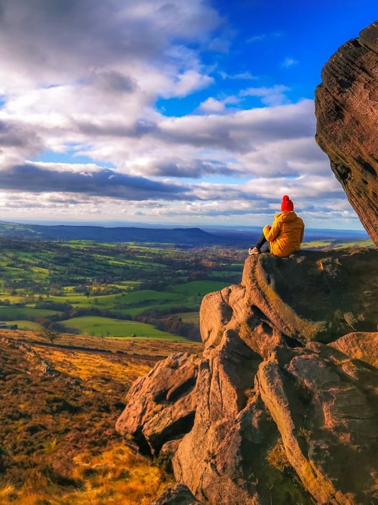 This may contain: a person sitting on top of a large rock next to a lush green valley under a cloudy blue sky