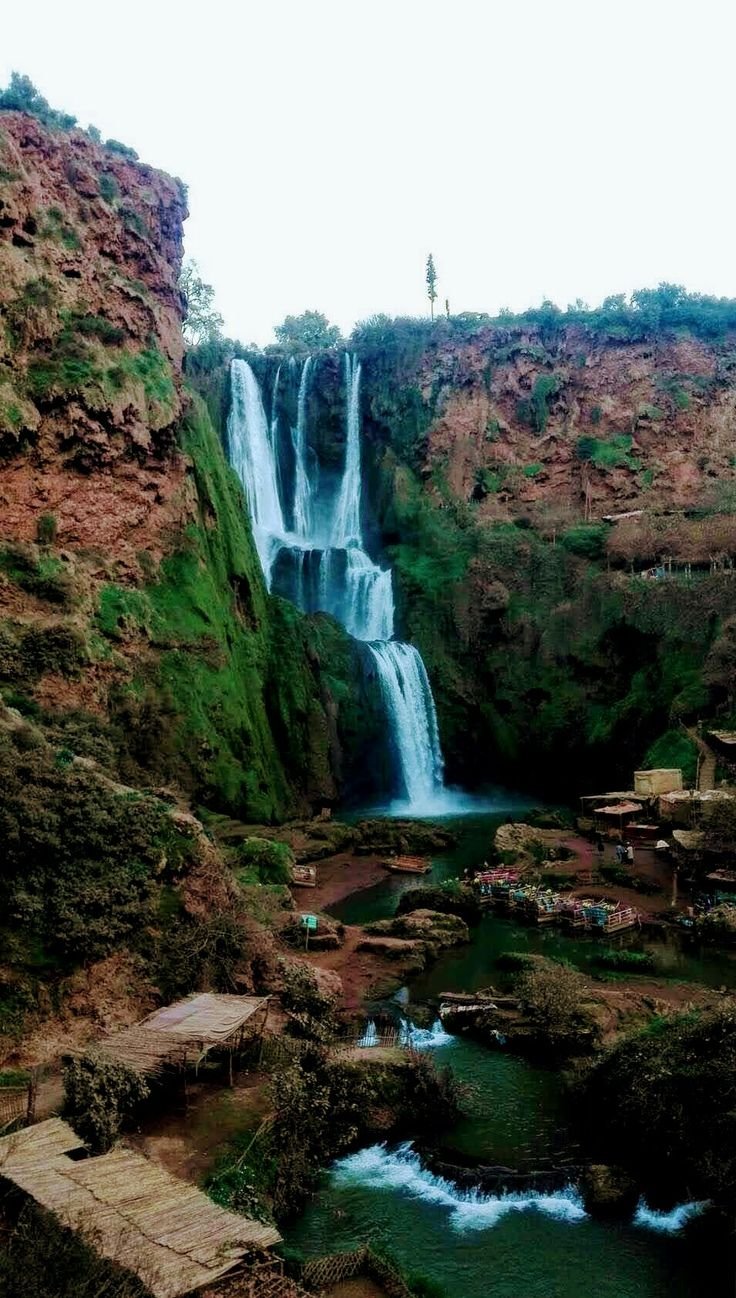 This may contain: a man standing at the base of a tall waterfall next to a body of water
