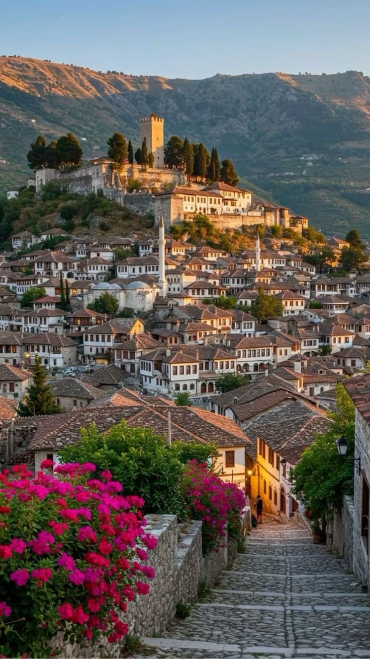 This may contain: an old town with lots of flowers growing on the steps leading up to it and mountains in the background