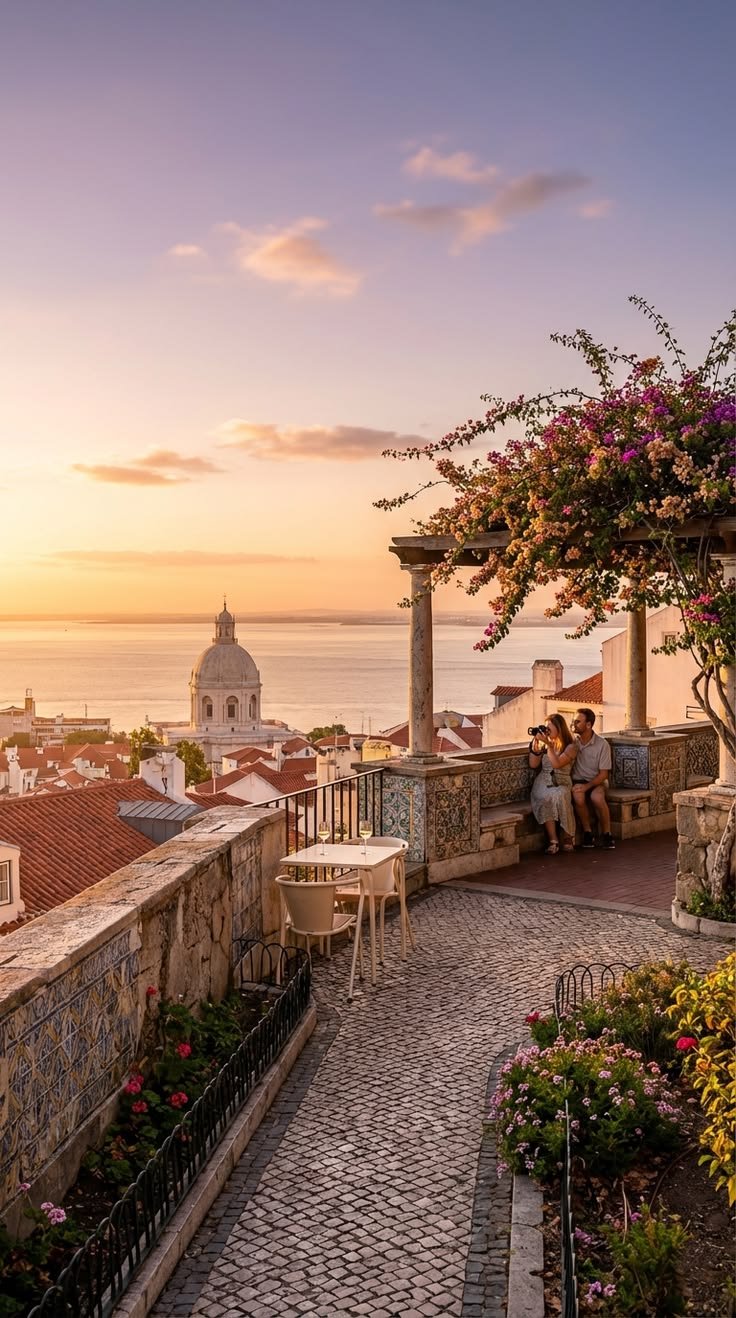 This may contain: two people are sitting on a bench overlooking the ocean and buildings at sunset, with pink flowers in the foreground